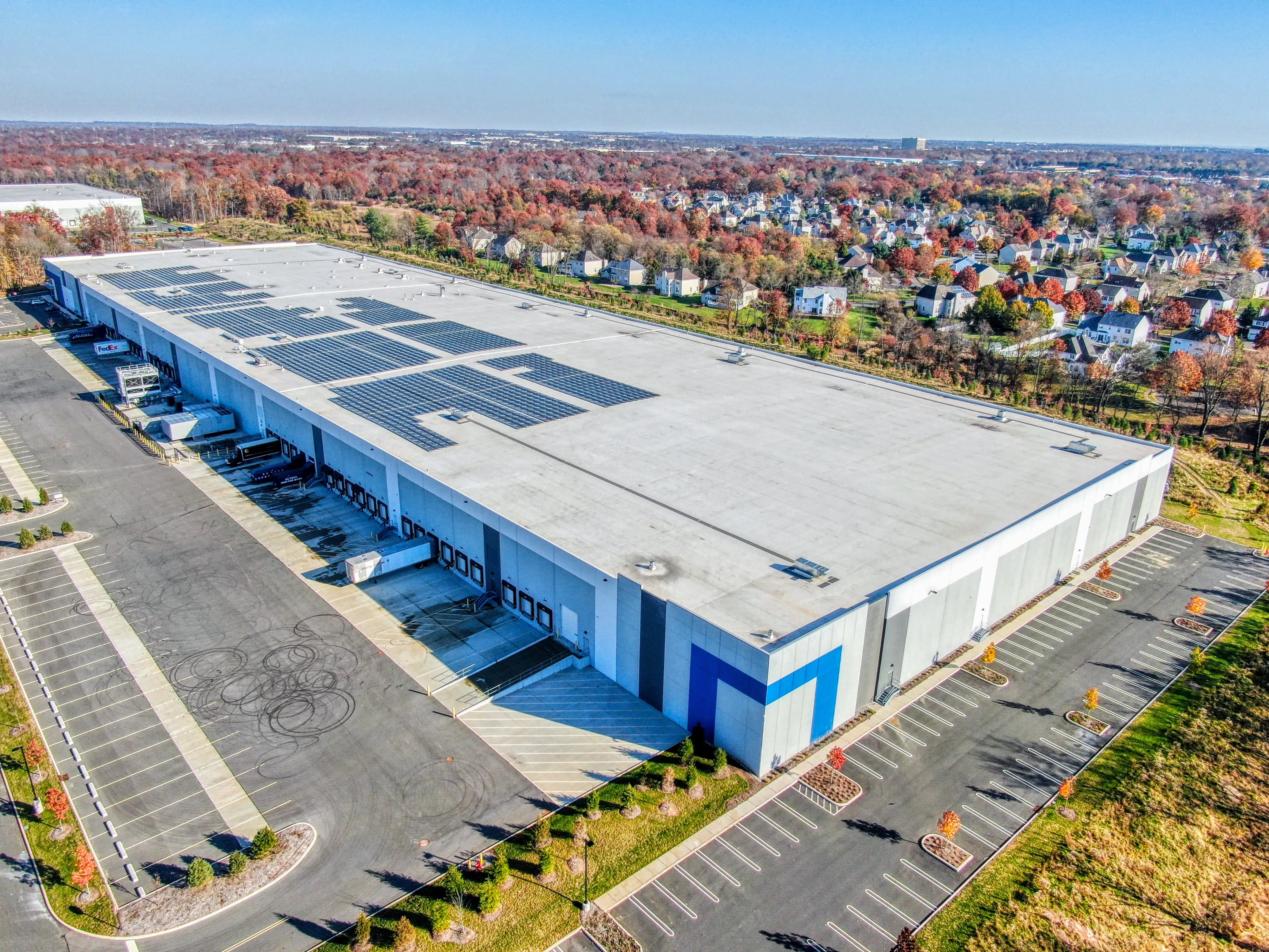 Aerial view of a large distribution center with a solar-panel roof and loading docks, autumn suburb in the background