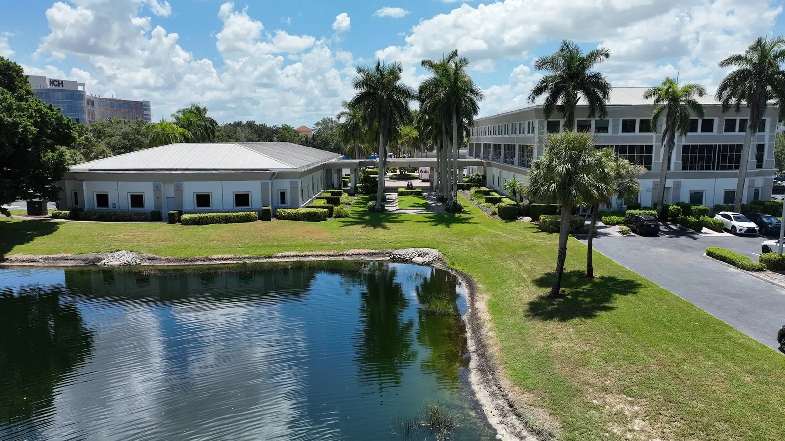 Drone footage of a Naples, Florida medical office campus, from the nearby intersection to the pond view, a push-in through the courtyard to the entrance, a top-down over the parking and roofs, and a closing rise over the fountain pond.